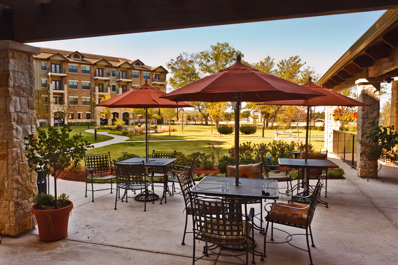 Outdoor seating area with metal tables and chairs under red umbrellas on a concrete patio, overlooking a landscaped garden with trees and a multi-story residential building in the background.