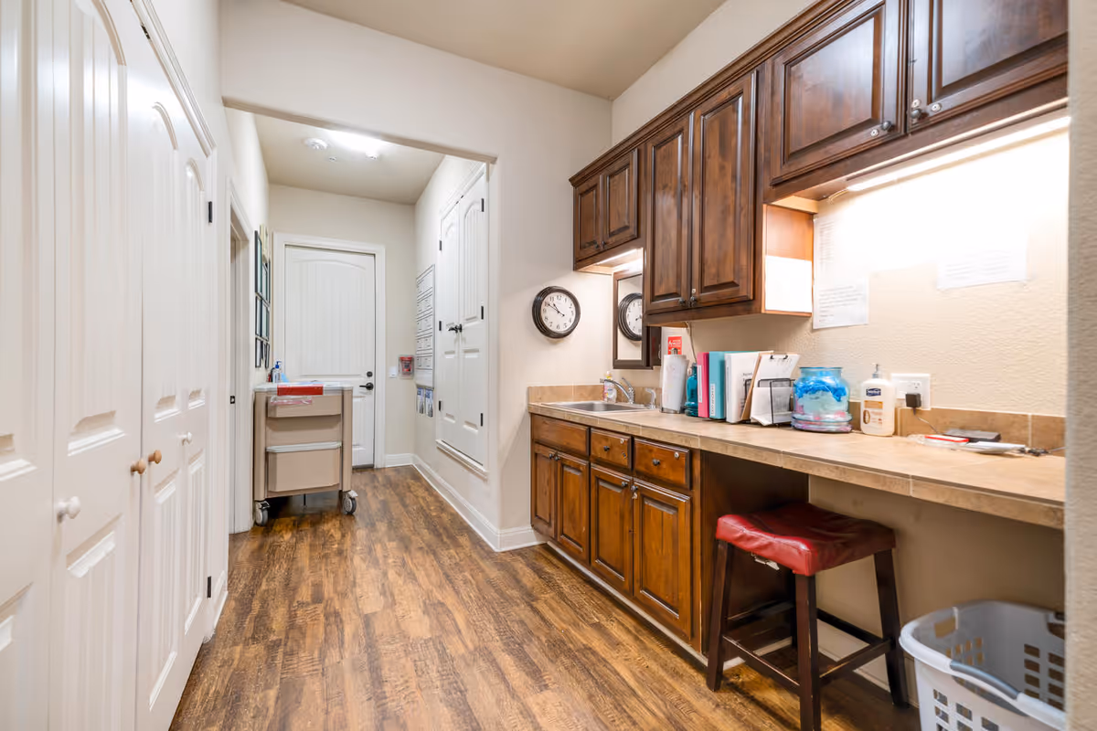 A hallway with wooden flooring and white walls featuring a countertop with dark wooden cabinets above and below. On the countertop are various items including books, a clock, a water dispenser, and hand sanitizer. There is a red cushioned stool under the counter and a laundry basket on the floor. At the end of the hallway is a door and a cart with drawers.