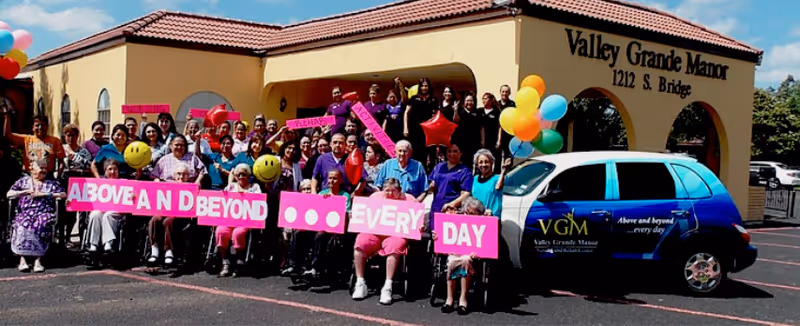 A large group of residents and staff pose with pink signs, balloons and a branded car in front of the Valley Grande Manor building.