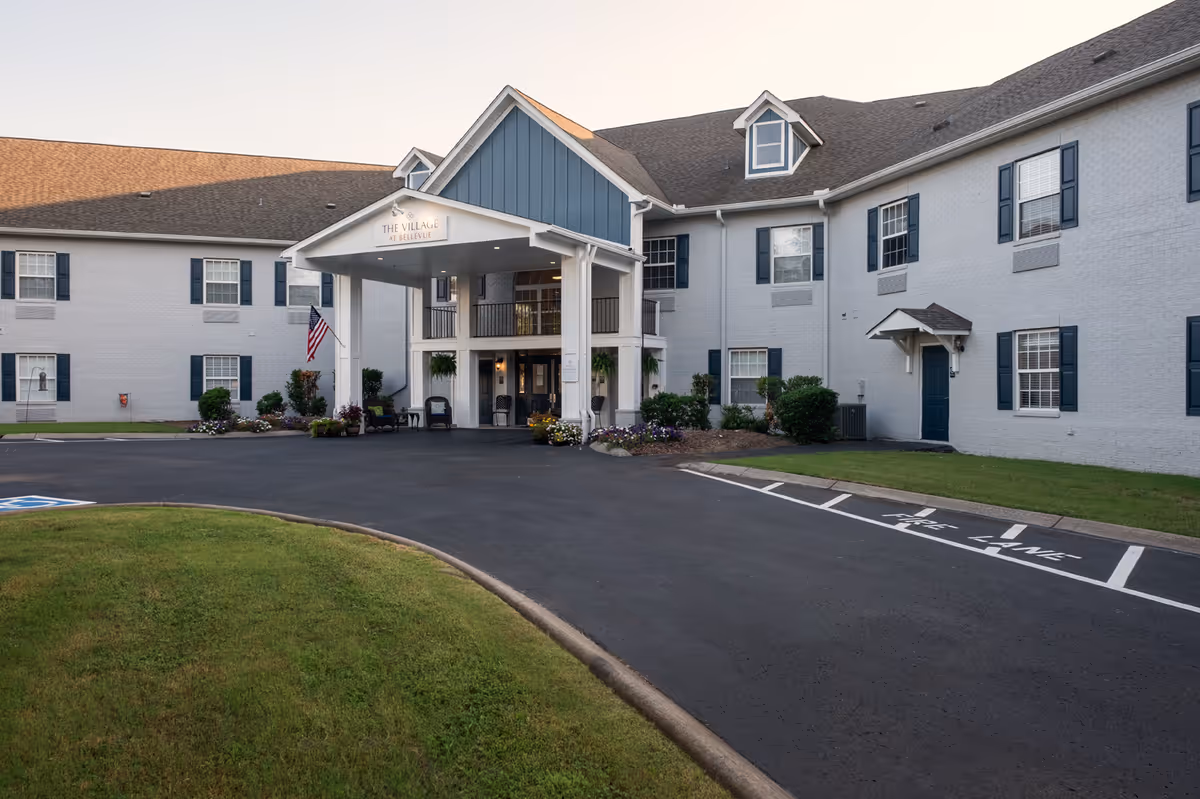 Exterior front view of The Village at Bellevue, a two-story assisted living facility with a covered entrance, multiple windows with blue shutters, an American flag, and landscaped greenery around the building.