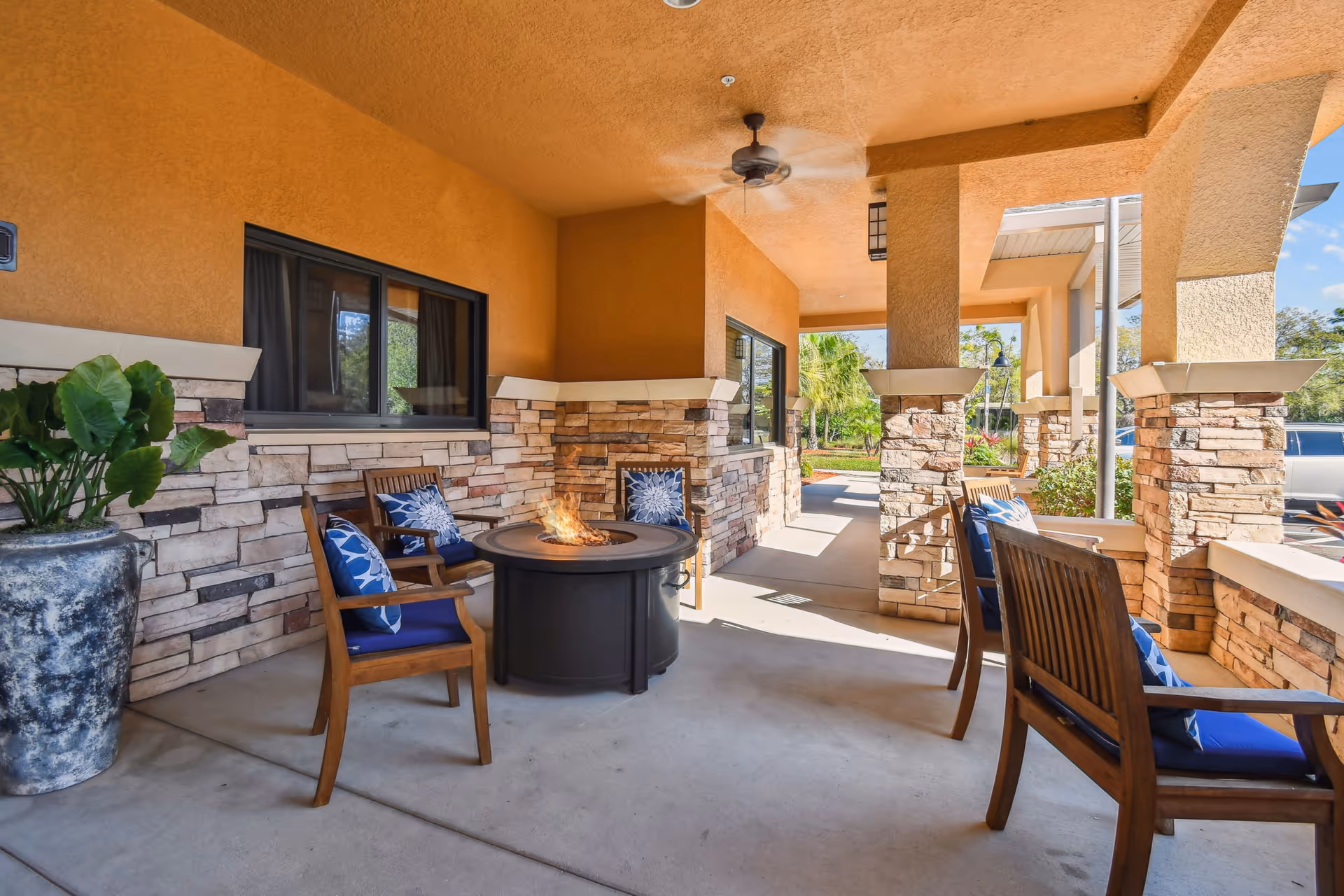 Covered outdoor patio area with stone pillars and walls, featuring wooden chairs with blue cushions arranged around a round fire pit table. A large potted plant is on the left side, and ceiling fans are mounted overhead. The area opens to a landscaped garden with palm trees and a clear blue sky.