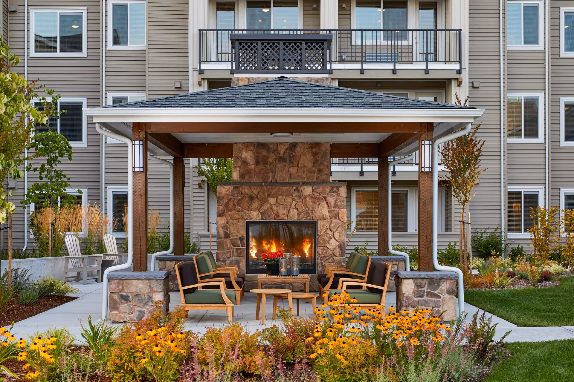 Covered outdoor seating area with a stone fireplace and chairs in front of a multi-story residential building surrounded by landscaped flowers.