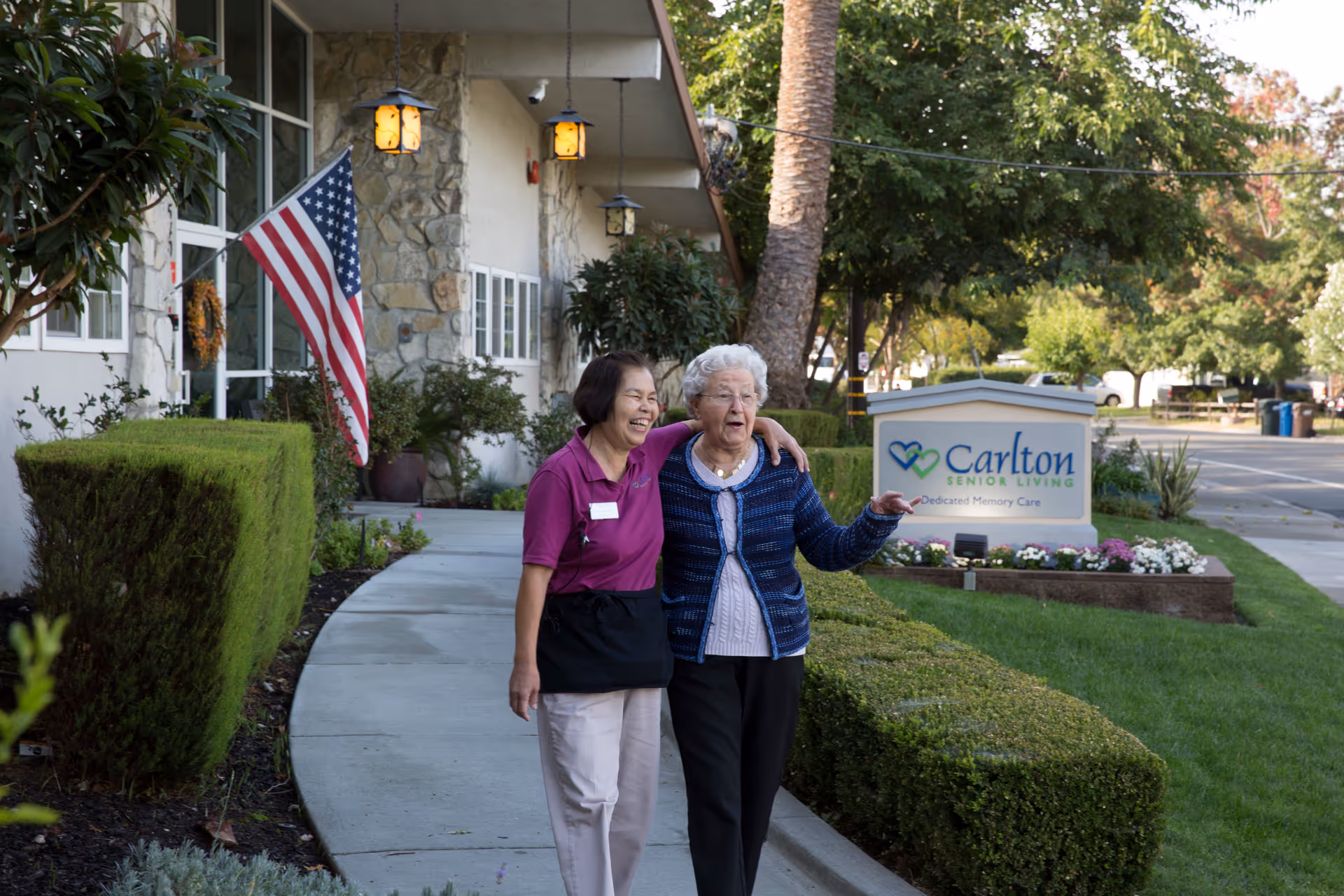 An elderly woman and a caregiver walking arm in arm along a sidewalk outside a senior living facility. The building has stone and white walls, with an American flag displayed near the entrance. There are trimmed bushes and trees along the walkway, and a sign that reads 'Carlton Senior Living Dedicated Memory Care' is visible in the background.