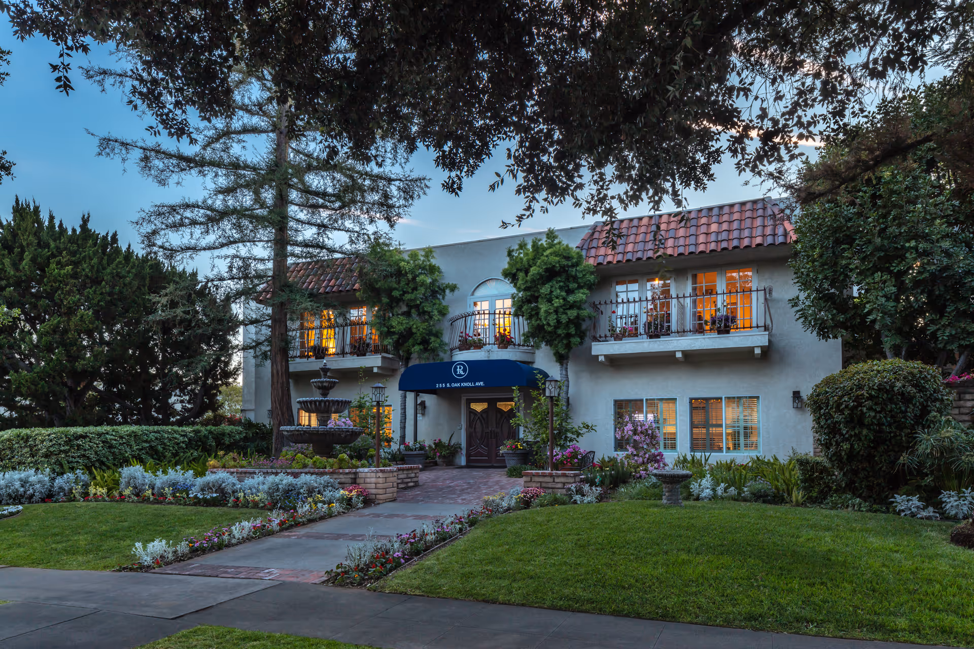 Exterior view of a two-story building with a tiled roof and balconies, surrounded by well-maintained gardens and trees. A pathway leads to the entrance, which has a blue awning with the letter 'R' and the address '255 S. OAK KNOLL AVE'. There is a multi-tiered fountain on the left side of the entrance.