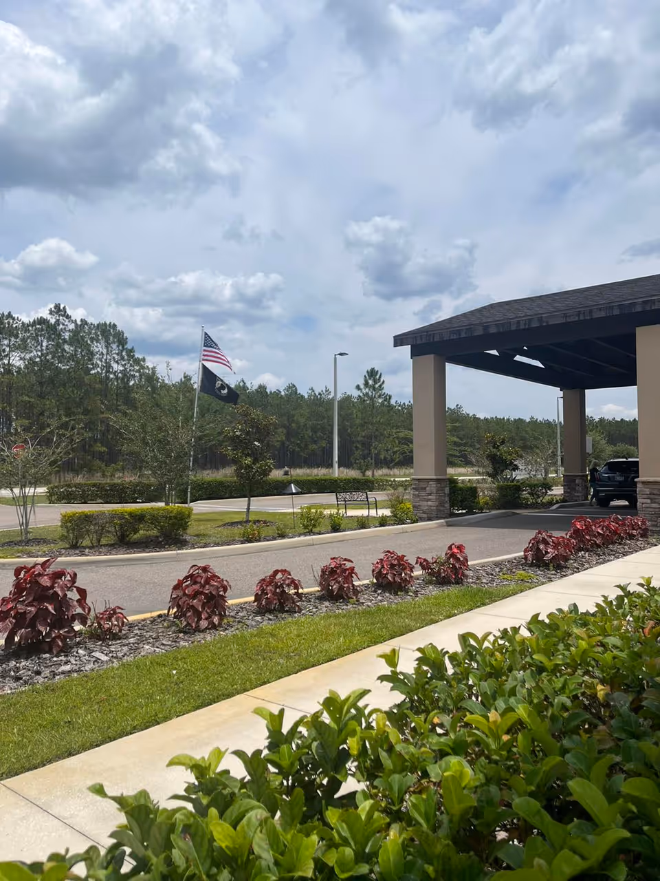 Outdoor view of the entrance area of a senior living facility with a covered drop-off area, landscaped bushes, a sidewalk, and two flags on flagpoles against a partly cloudy sky.