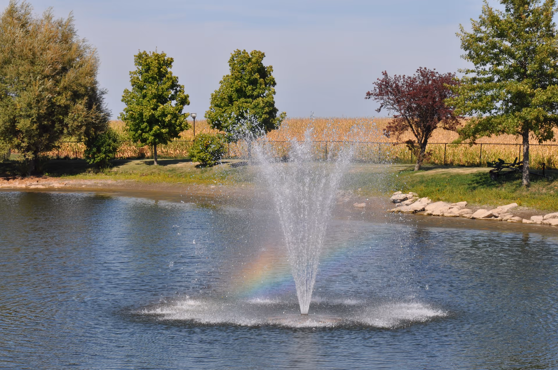 A water fountain spraying water upwards in the middle of a pond with a faint rainbow visible in the mist. Trees and a grassy area surround the pond under a clear sky.