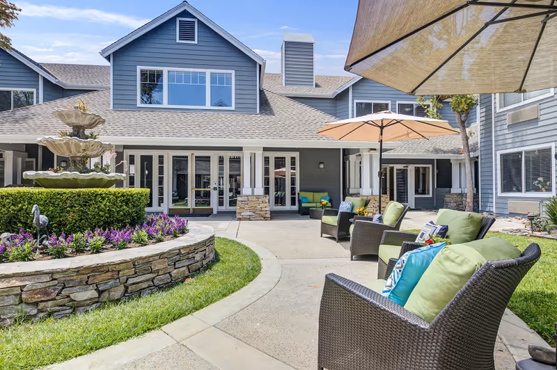 Outdoor courtyard area of a senior living facility with a stone fountain surrounded by purple flowers and greenery. There are several cushioned wicker chairs with colorful pillows arranged under large umbrellas on a paved patio. The building in the background is two stories with gray siding, white trim, and multiple windows and doors leading to the courtyard.