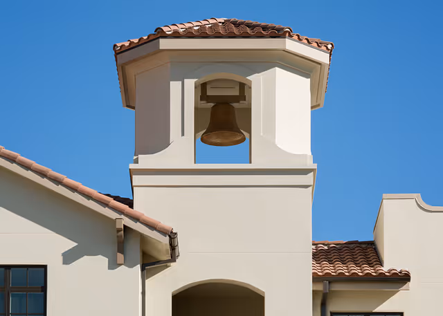 Light-colored Mediterranean-style building tower with a large bell under a tiled roof against a clear blue sky.