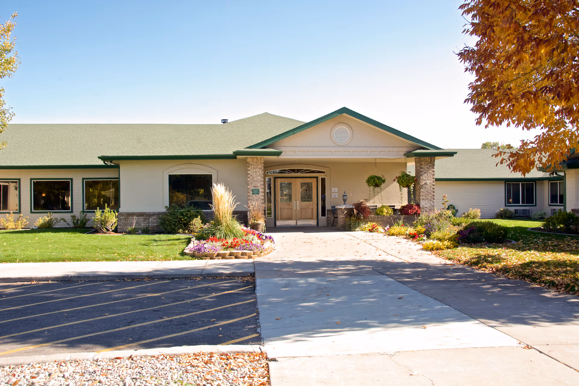 Front exterior view of Brookdale Chubbuck facility with a green roof, beige walls, large windows, and a covered entrance. There are landscaped flower beds with colorful flowers and green grass on either side of the entrance. Trees with autumn-colored leaves frame the scene.