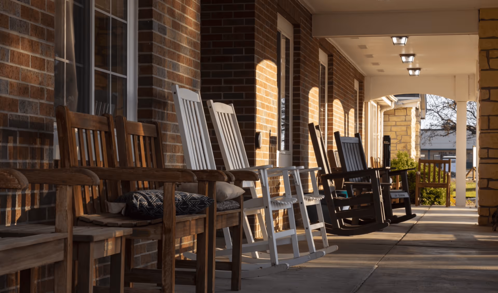 A covered brick porch lined with wooden and white rocking chairs bathed in warm sunlight.