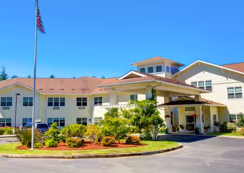 Exterior view of a two-story senior living facility building with beige siding and a red roof. There is a covered entrance with columns, a landscaped garden with bushes and small trees, and an American flag on a flagpole in front of the building under a clear blue sky.