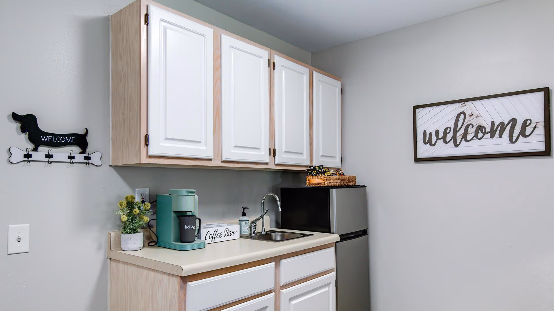 Small kitchenette area with light wood and white cabinets, a countertop with a green coffee maker, a small sink, a soap dispenser, and a mini refrigerator. On the wall above the counter is a framed sign that says 'welcome' and a decorative black dog-shaped hook rack with the word 'WELCOME'.
