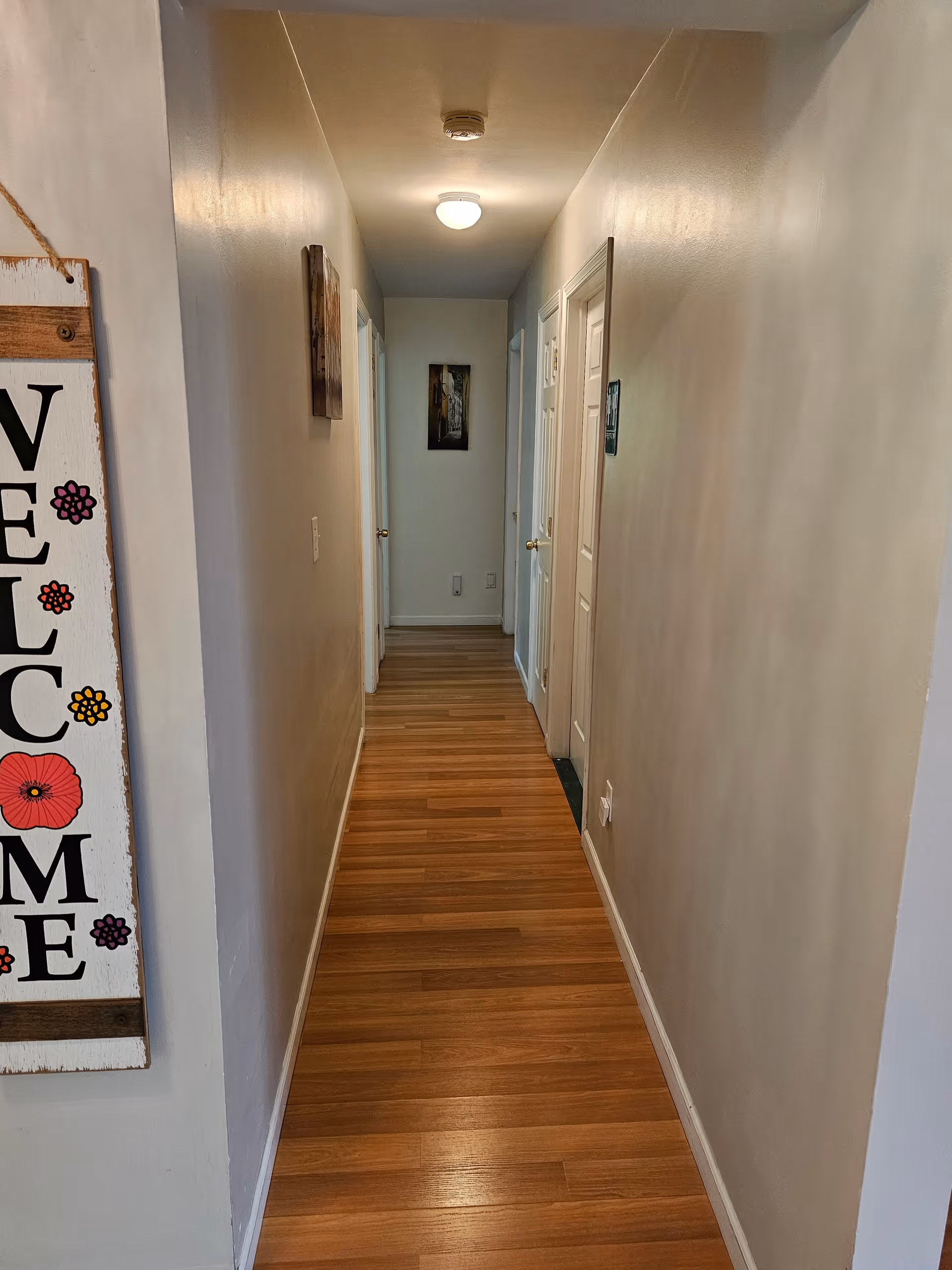 A narrow hallway with wooden flooring and light-colored walls. There are several closed white doors along the right side and one door on the left. A ceiling light fixture illuminates the hallway. On the left wall near the foreground, there is a decorative sign with the word 'WELCOME' and colorful flowers.