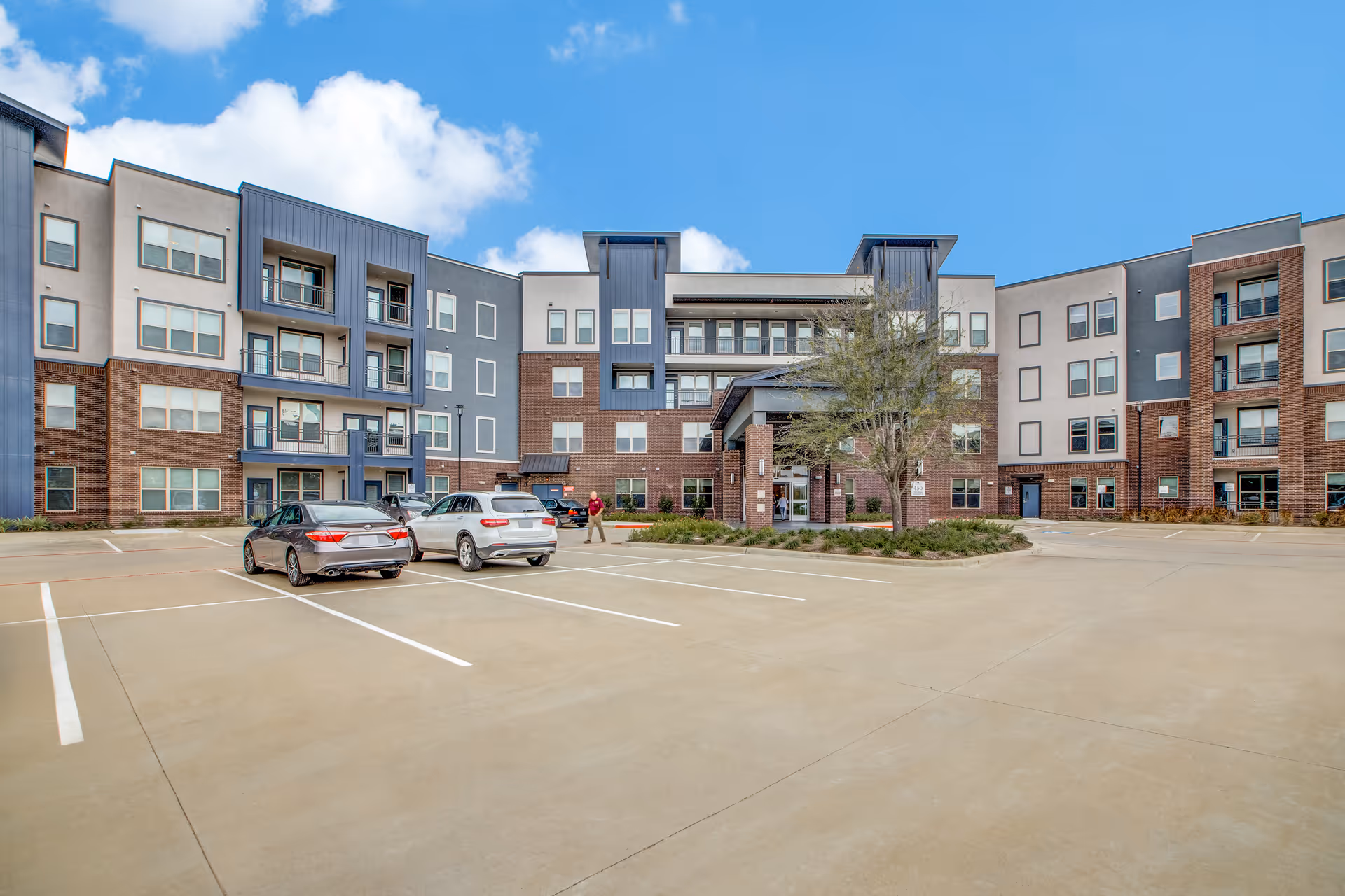 Exterior view of a modern multi-story senior living facility with a large parking lot in front, two cars parked, and a person walking near the entrance under a blue sky with some clouds.