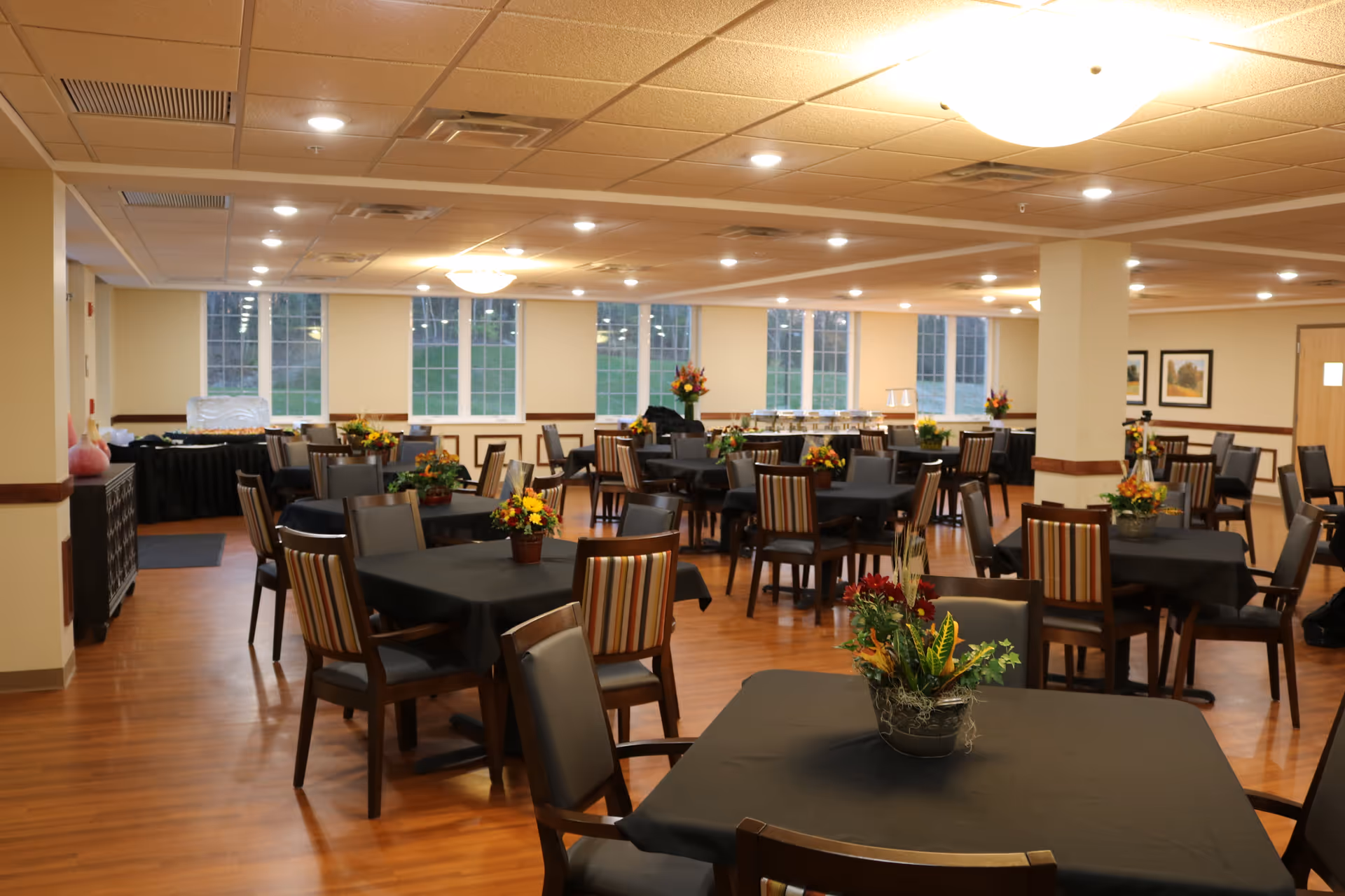 A spacious dining room with multiple tables covered in black tablecloths and decorated with flower arrangements. The room has large windows letting in natural light, wooden flooring, and a ceiling with recessed lighting. There are chairs with striped and solid backs around the tables, and buffet tables are set up along the walls.