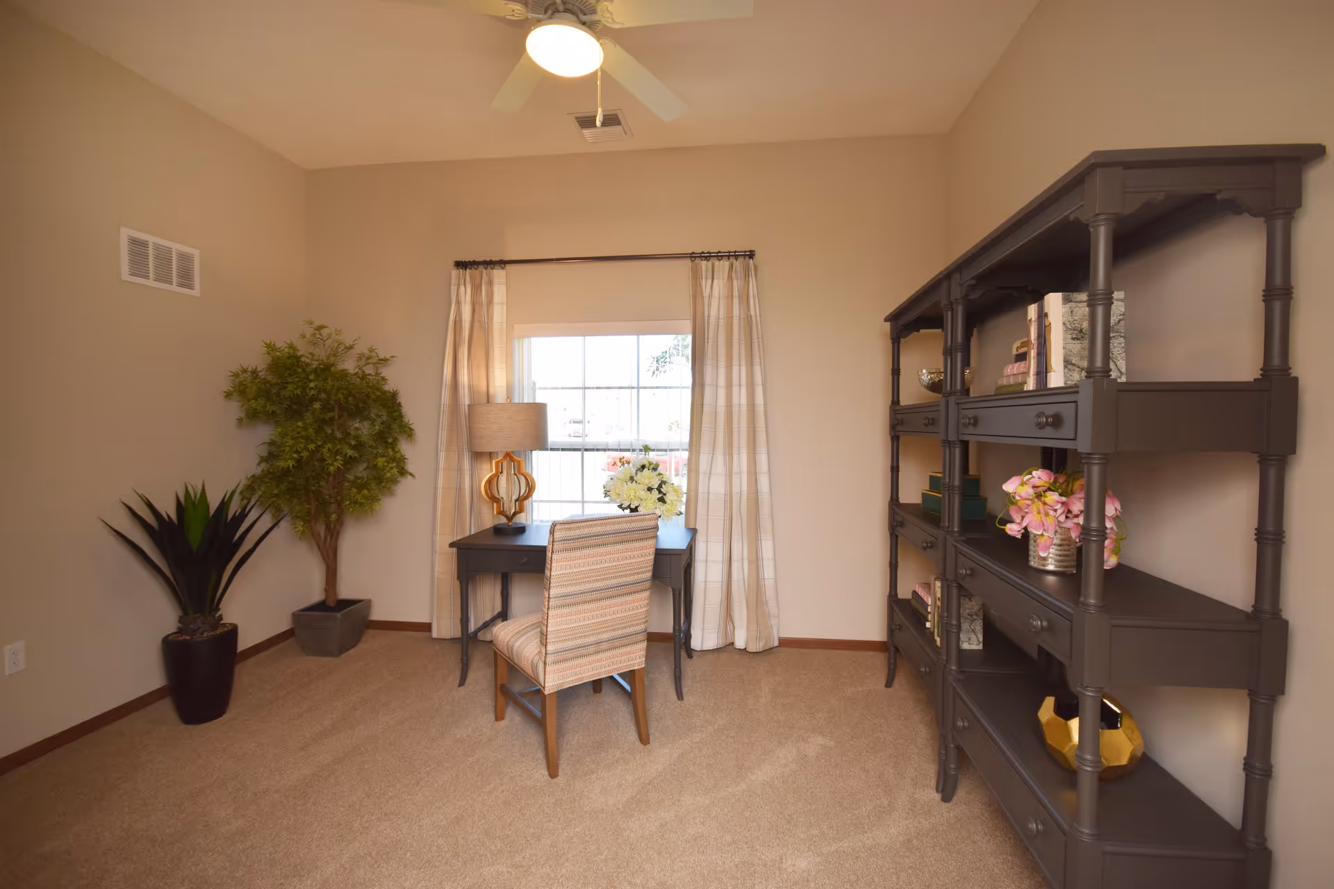 A cozy interior room with beige walls and carpet, featuring a small desk with a striped chair in front of a window with light-colored curtains. A table lamp and a vase with flowers are on the desk. To the right, there is a dark wooden shelving unit with decorative items and books. Two potted plants are placed in the corner to the left.