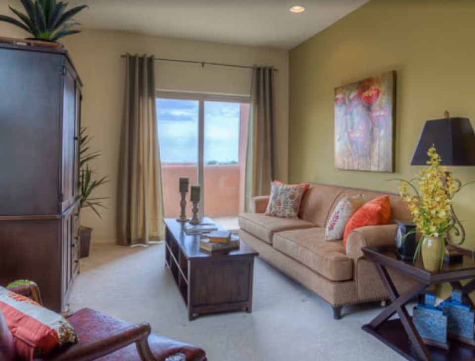 Bright living room with a beige sofa, coffee table, armchair, artwork on the wall, and a sliding glass door to a balcony.