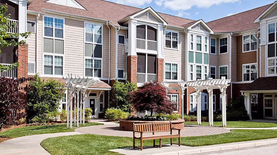 Outdoor courtyard area of a senior living facility with a wooden bench, a circular brick planter with a small tree, white pergolas, and a multi-story building with large windows in the background.