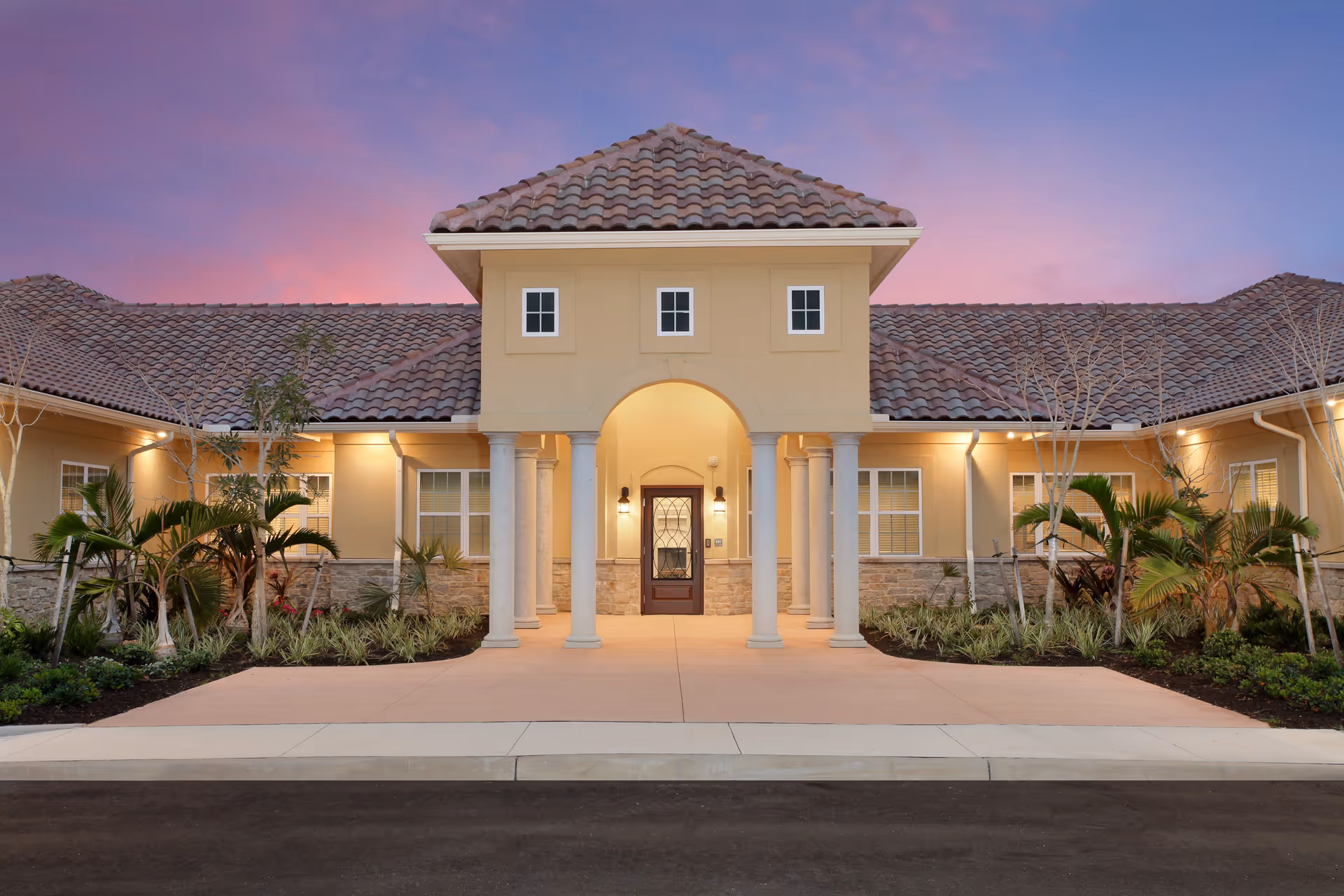 Front exterior view of a single-story building with a tiled roof, beige walls, and a central entrance featuring an archway supported by columns. The entrance is illuminated by wall-mounted lights, and there are landscaped plants and shrubs on either side of the walkway leading to the door. The sky is a gradient of purple and pink, indicating dusk or dawn.