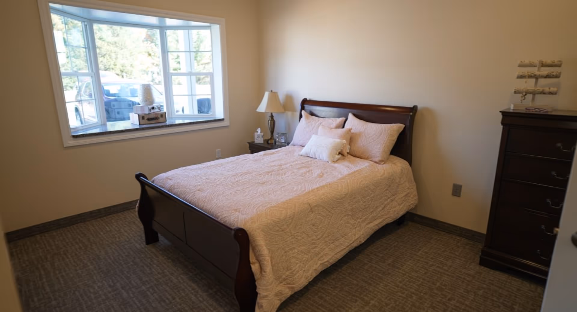 A bedroom with a wooden bed frame and a light pink quilted bedspread with matching pillows. There is a nightstand with a lamp and tissue box next to the bed, a window with a view of trees and a parked car outside, and a dark wooden dresser with a jewelry stand on top.