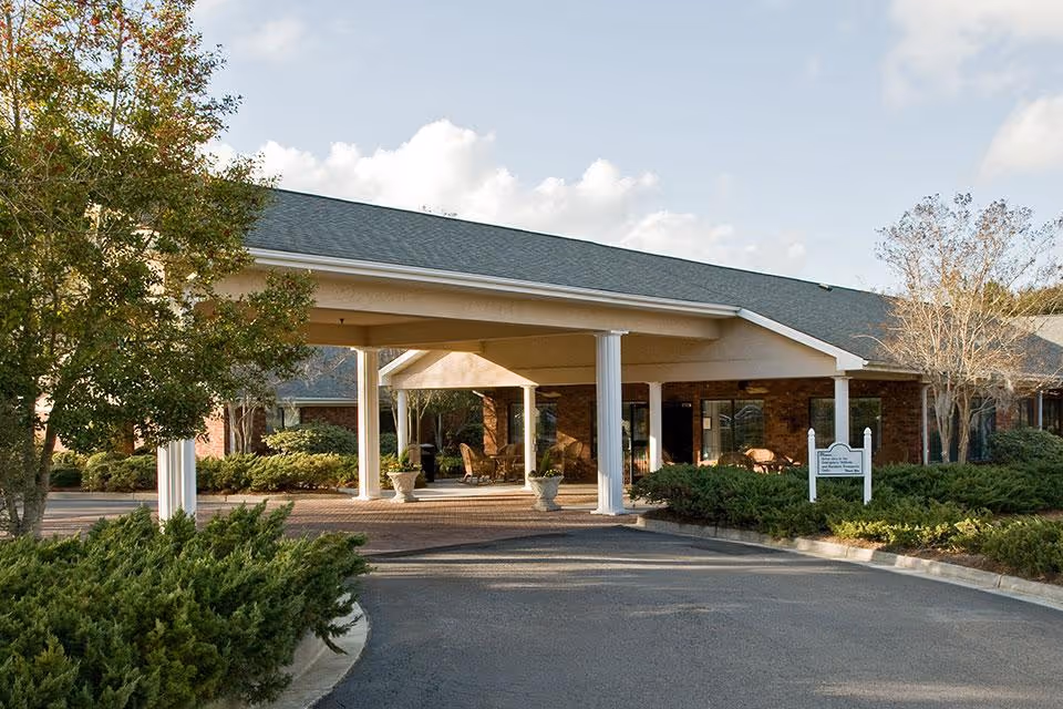 Front entrance of a single-story senior living facility with a covered drive-through portico, white columns, brick facade and surrounding landscaping.