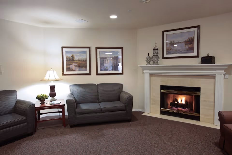 A cozy living room area with a lit fireplace, two gray upholstered armchairs, a matching loveseat, a wooden side table with a lamp and a small plant, and three framed landscape paintings on the walls.