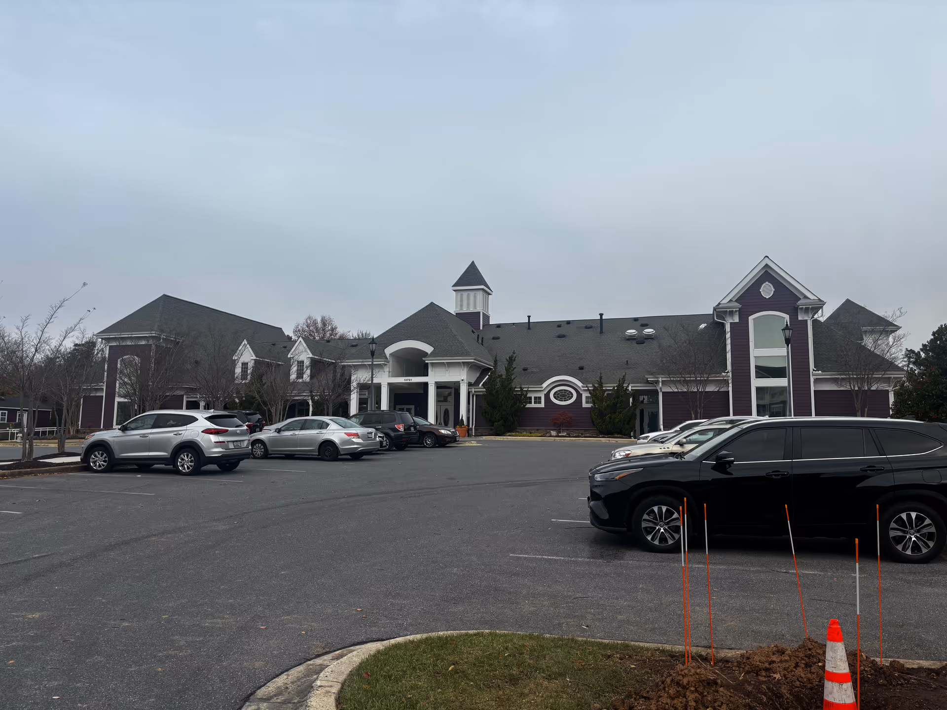 Large purple-and-white senior living building with a central entrance and several parked cars in the front parking lot under an overcast sky.