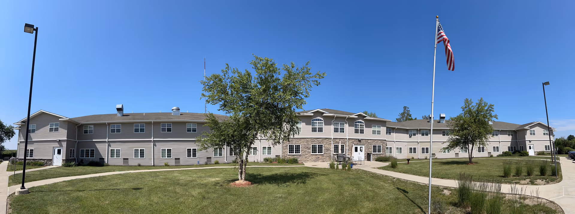 Wide panoramic view of a two-story senior living facility building with beige siding and stone accents, surrounded by a well-maintained lawn with a few trees, a flagpole with an American flag, and clear blue sky above.