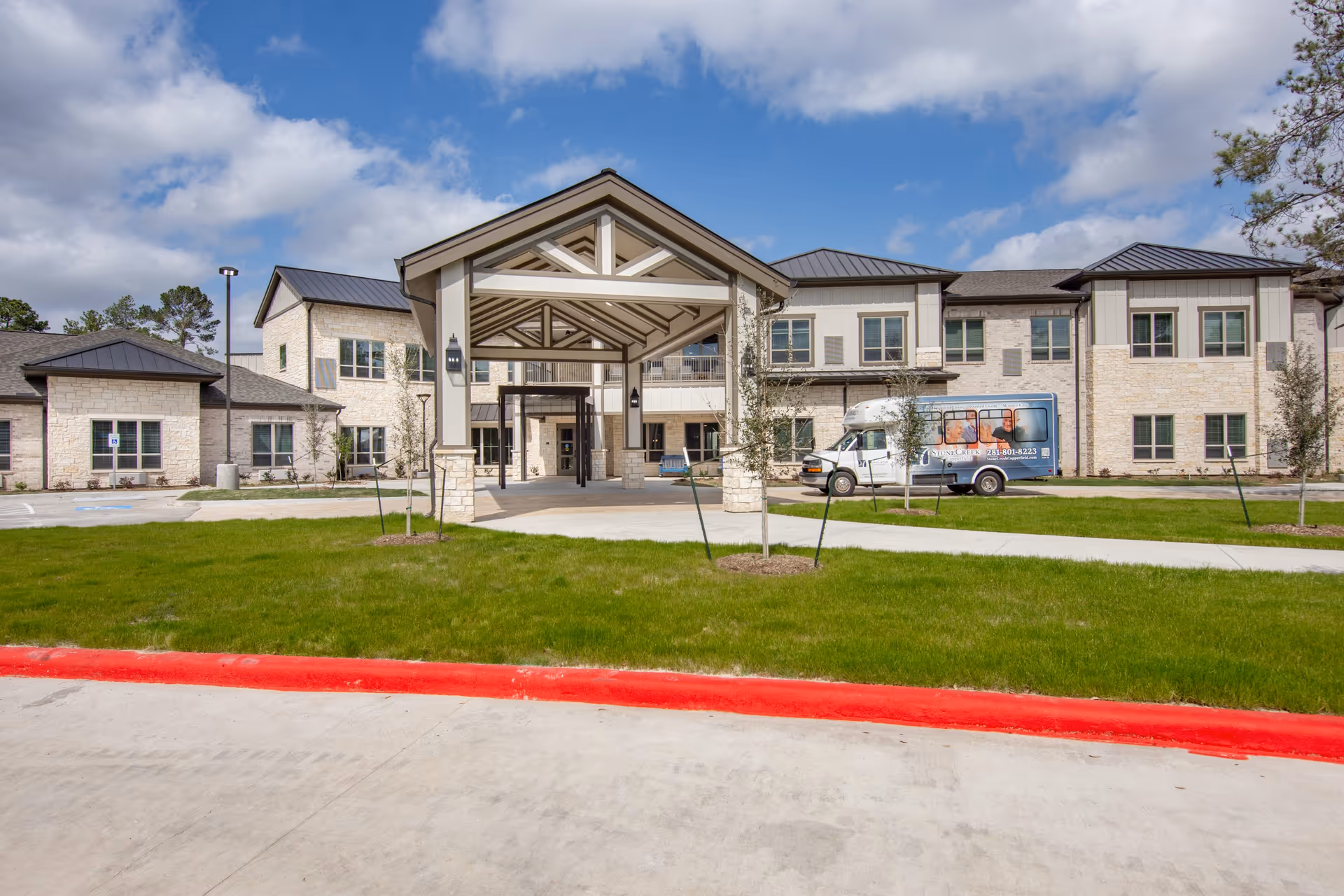 Exterior view of StoneCreek of Copperfield Senior Living facility showing a two-story building with stone and siding facade, a covered entrance with pillars, a green lawn, and a shuttle van parked near the entrance under a partly cloudy sky.