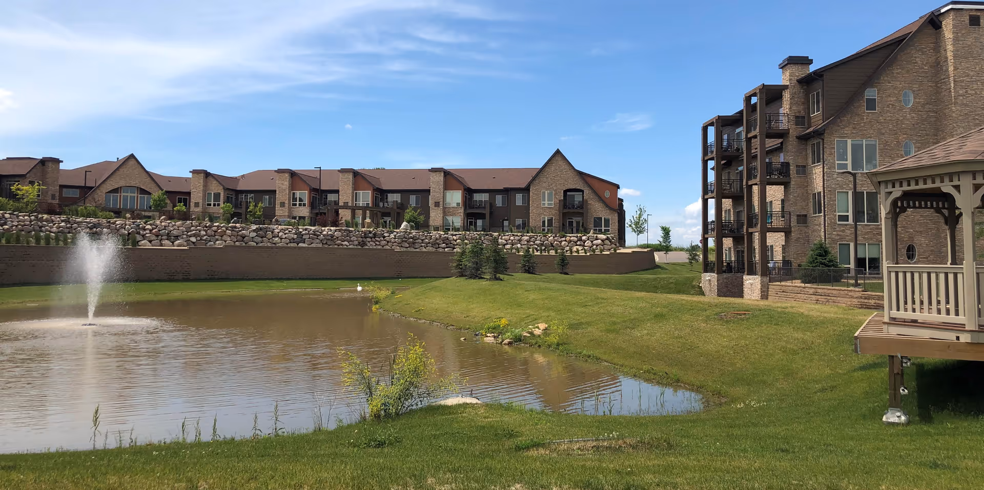 A pond with a fountain, grassy lawn and gazebo in front of multi-story senior living buildings under a blue sky.