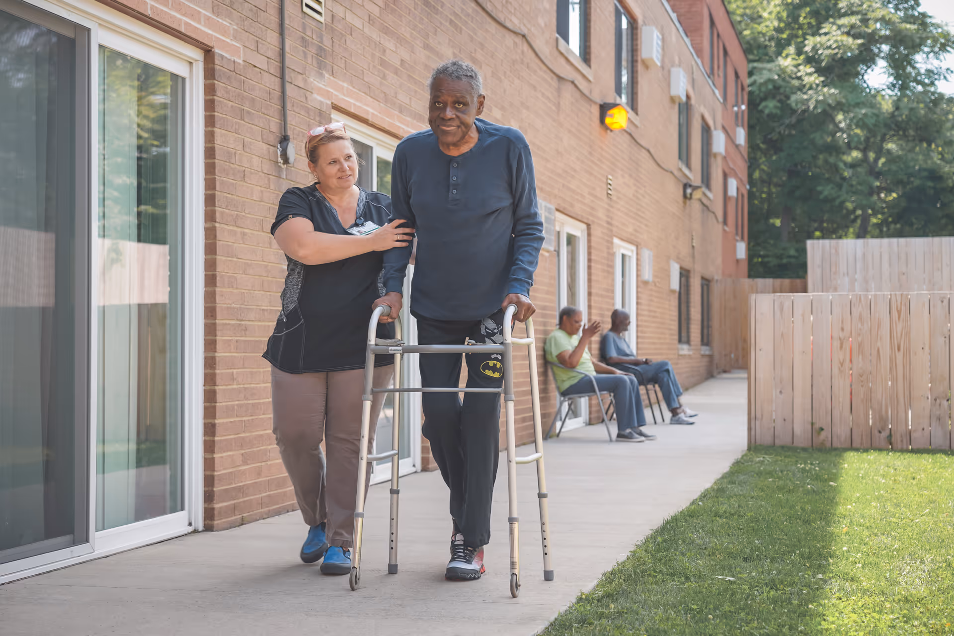 An elderly man using a walker is assisted by a caregiver as they walk along a sidewalk outside a brick building. Two other elderly men are seated on chairs further down the sidewalk near a wooden fence, with grass and trees visible on the right side.