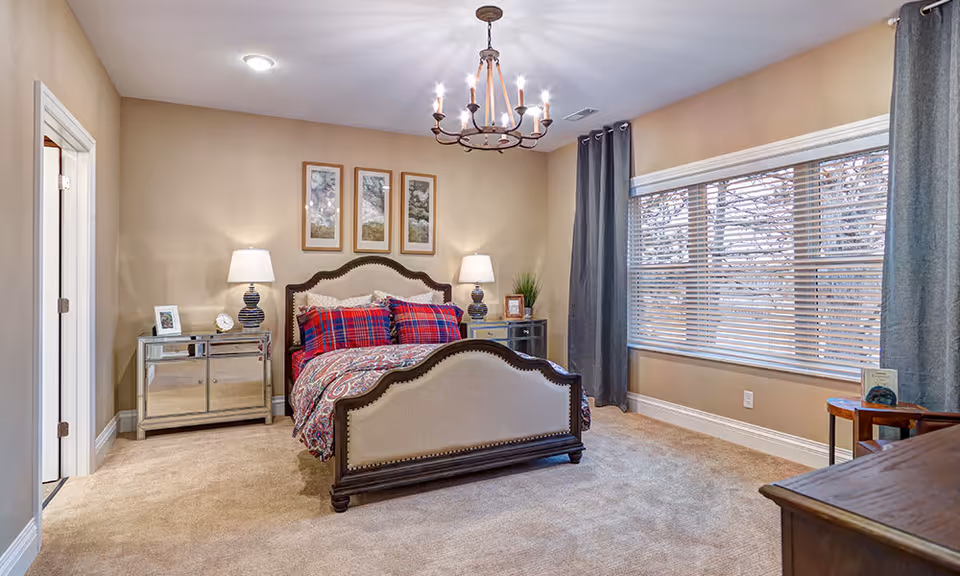 A cozy bedroom with a large upholstered bed featuring red and blue plaid bedding. There are two mirrored nightstands on either side of the bed, each with a lamp. Above the bed, three framed pictures hang on the beige wall. A large window with gray curtains allows natural light into the room. The floor is carpeted in a light beige color, and a wooden dresser is partially visible in the foreground.