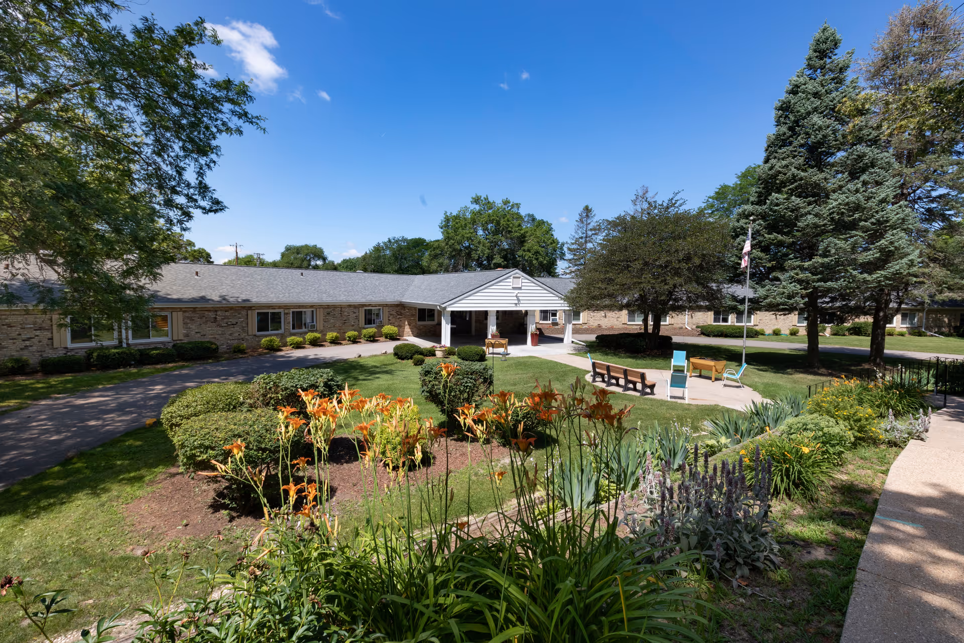 Exterior view of Madison Health and Rehabilitation Center showing a single-story building with a covered entrance, surrounded by a well-maintained garden with orange flowers, green shrubs, trees, benches, and a flagpole under a clear blue sky.