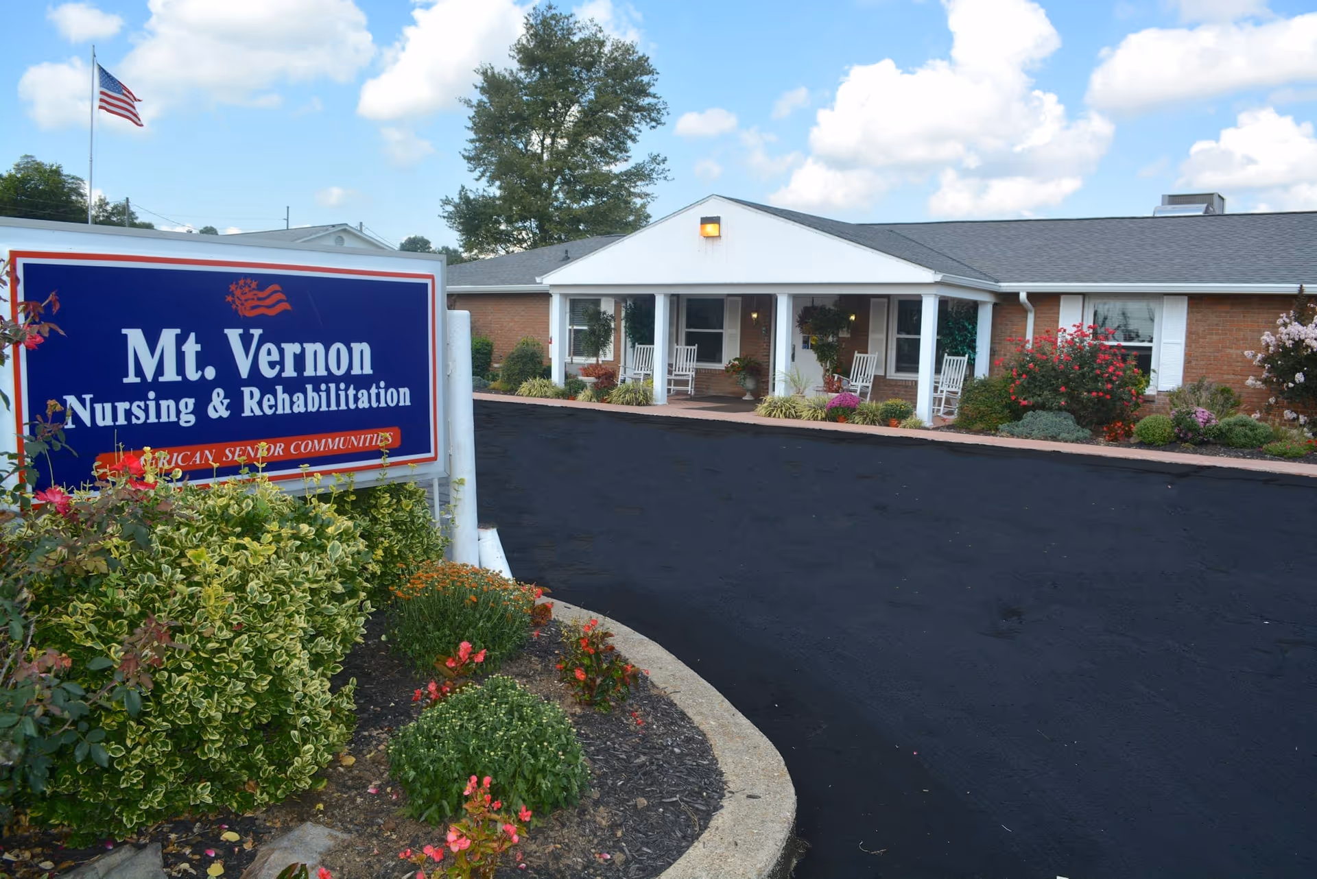 Front entrance of Mt. Vernon Nursing & Rehabilitation with its sign, landscaped flowerbeds, driveway, and a low brick building with a porch.