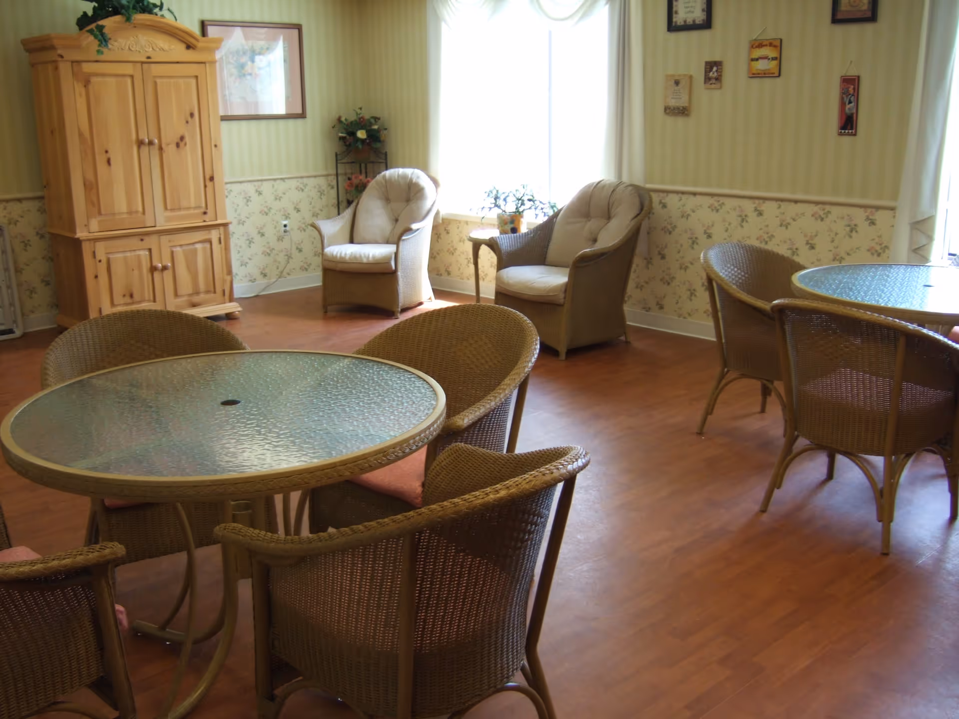 Bright common room with round glass-top tables, wicker chairs, and two cushioned armchairs by a window.