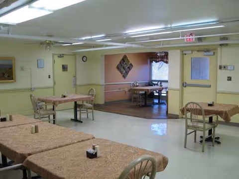 Sunlit dining room in a senior living facility with multiple brown tablecloth-covered tables and chairs.