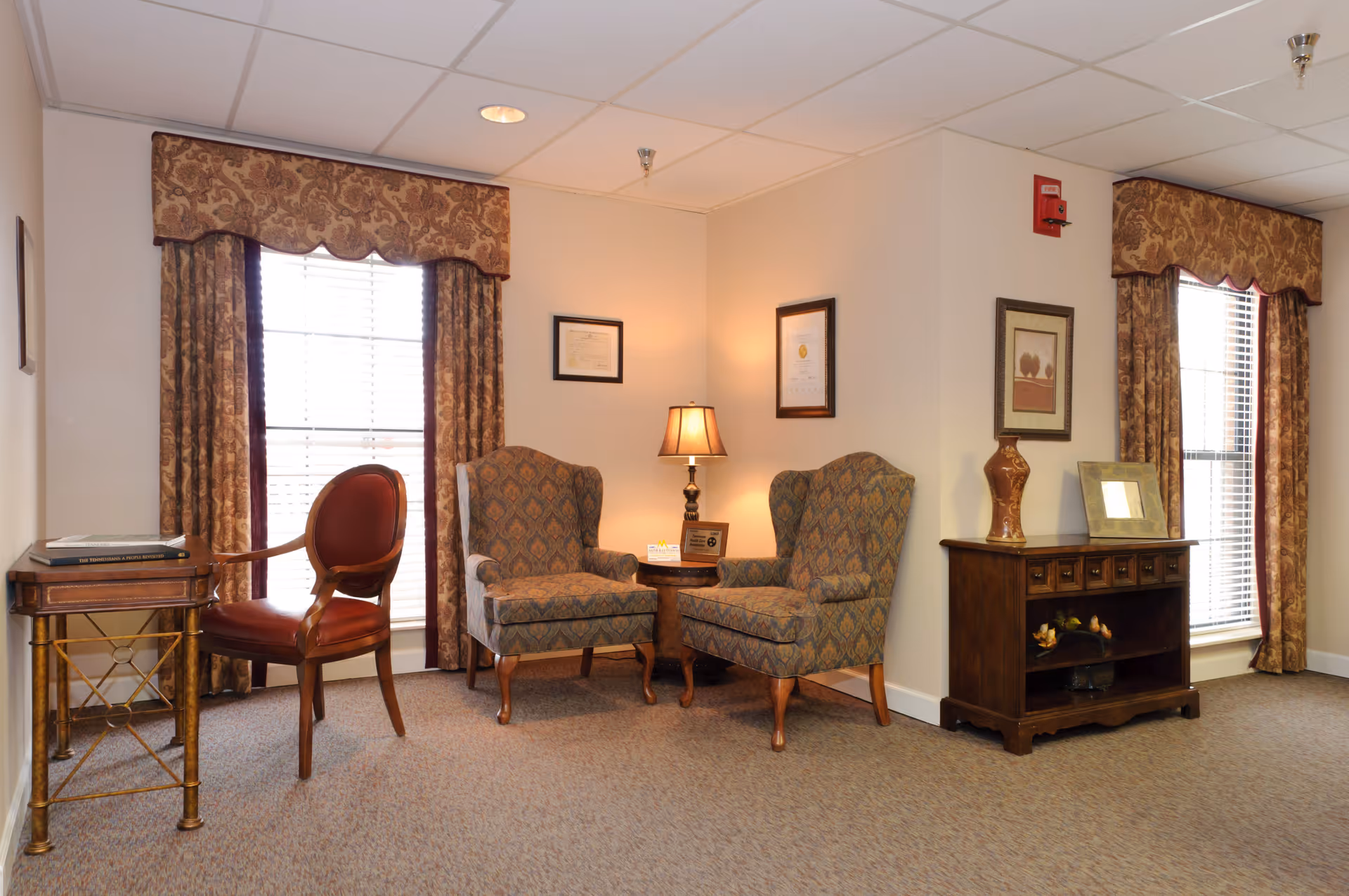 A cozy sitting area in a senior living facility with two patterned armchairs around a small wooden table with a lamp. There are two large windows with brown patterned curtains, a wooden side table with a red cushioned chair, and a wooden cabinet with decorative items and framed pictures on the walls.