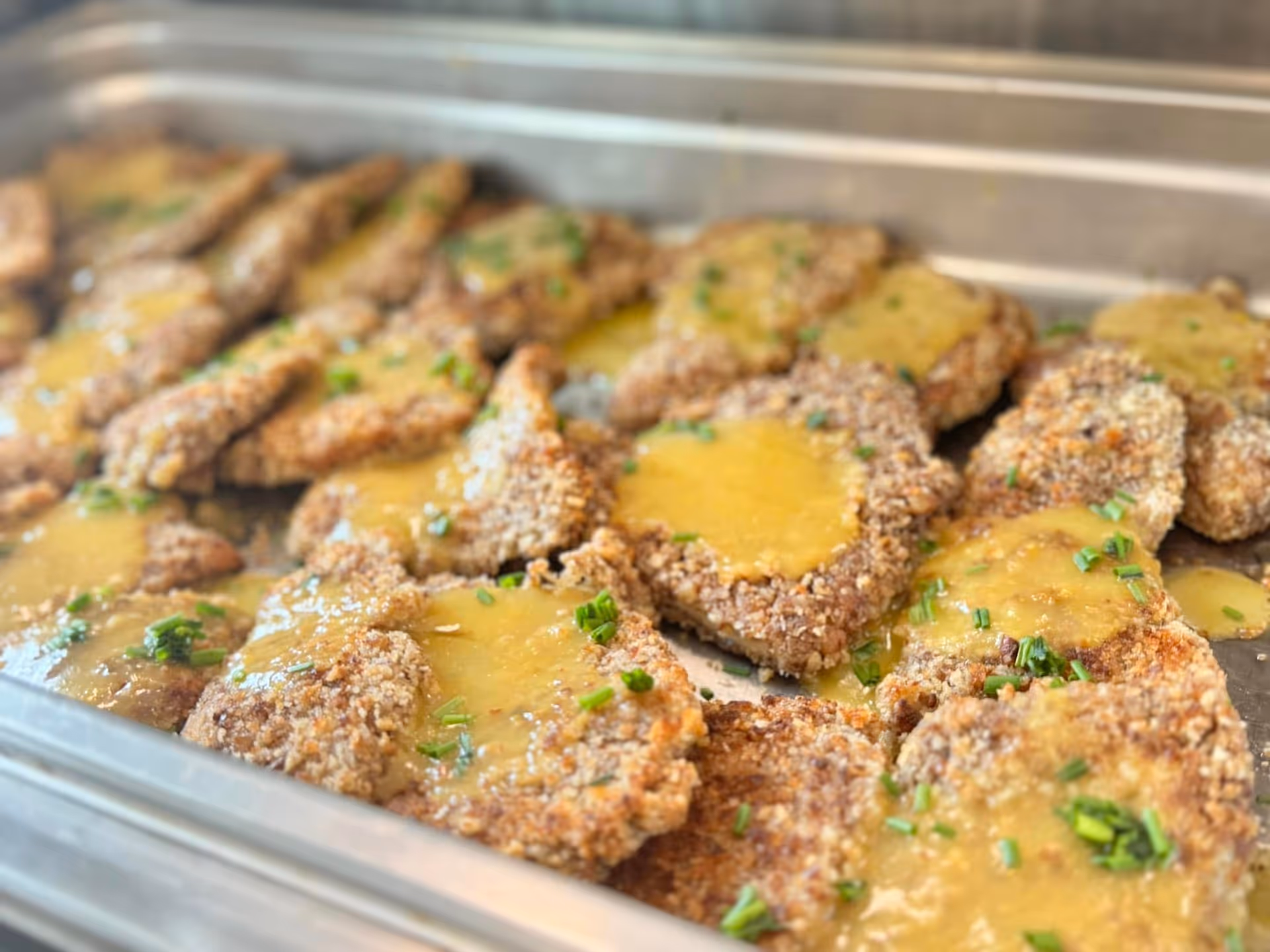 Breaded cutlets topped with a yellow sauce and chopped chives arranged in a metal serving tray.