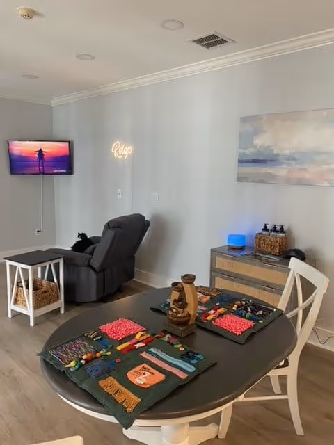 A cozy living area with a round table set with colorful textured mats and sensory items. In the background, there is a gray recliner chair facing a wall-mounted TV displaying a sunset scene. A small white side table with a basket underneath is next to the chair. On the wall, a neon sign reads 'Relax' and a large abstract painting hangs above a wooden cabinet with various items on top.