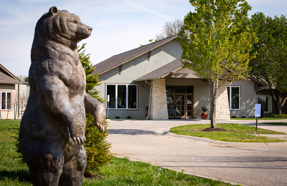 Outdoor view of a senior living facility entrance with a large bear statue in the foreground, a paved driveway, green grass, and trees surrounding the building.