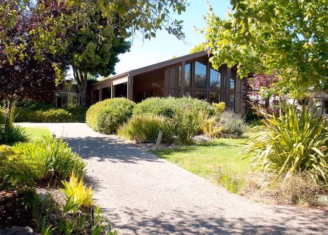 Sunlit brown single-story building with large windows set behind a paved walkway and surrounded by trees, shrubs, and landscaping.