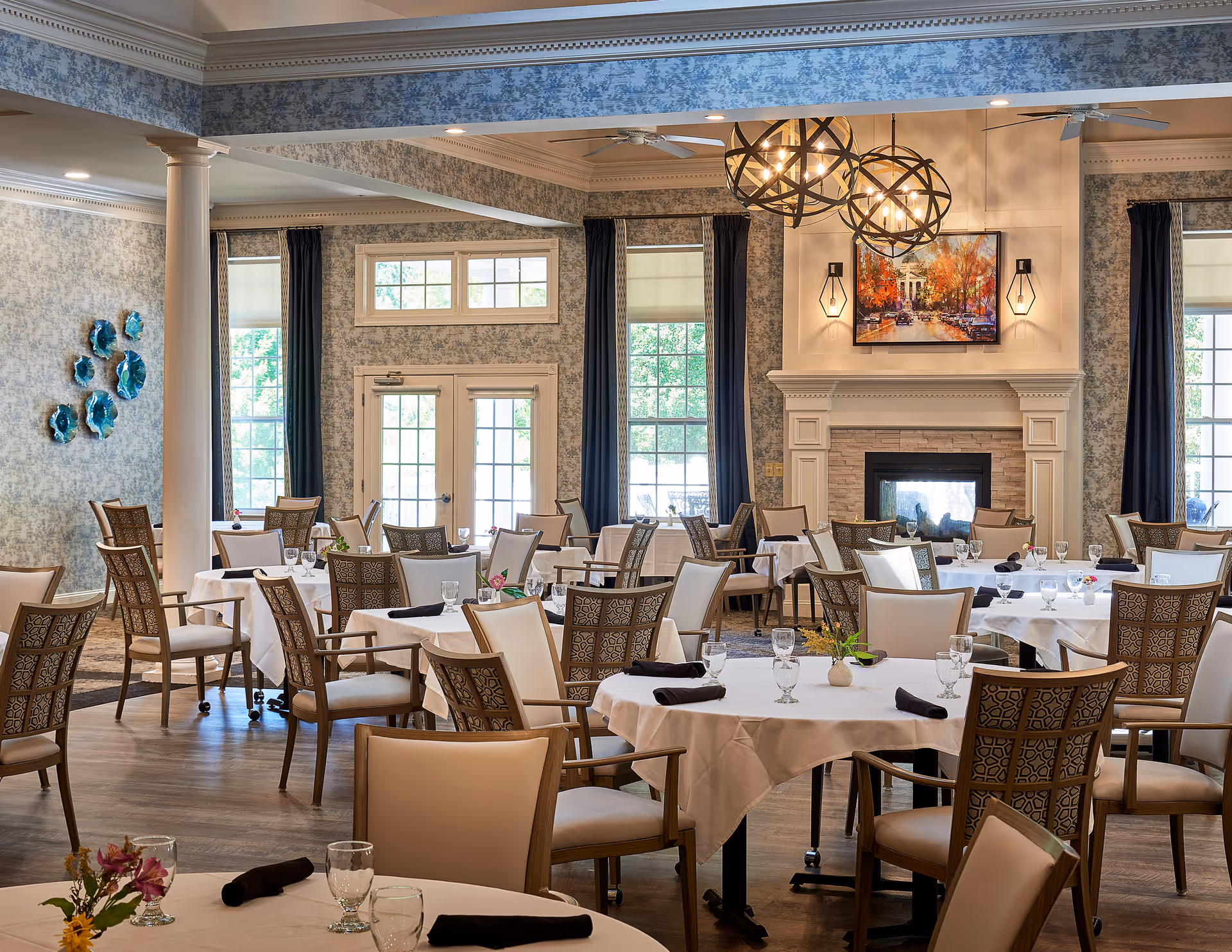 A spacious dining room with multiple round tables covered in white tablecloths, each set with glassware and black napkins. The room features elegant chairs with patterned backs, large windows with dark curtains, a fireplace with a colorful painting above it, and modern spherical chandeliers hanging from the ceiling. The walls have a textured blue and white design, and there are decorative blue wall art pieces on one side.