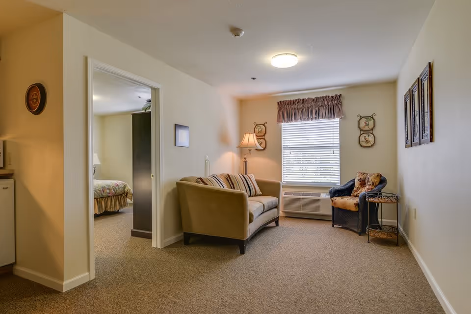 A cozy living room area in a senior living facility with a beige sofa, a striped cushion, a wicker armchair with floral cushions, a floor lamp, and a window with blinds and a valance. The room has beige walls and carpeted floor. A doorway leads to a bedroom with a bed and a dark wardrobe visible.