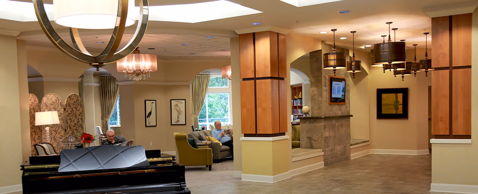 A spacious and well-lit senior living facility common area with comfortable seating including armchairs and sofas. Two elderly men are seated and engaged in reading or conversation. The room features warm beige walls, large windows with curtains, framed bird artwork, a decorative room divider, and modern light fixtures including a large circular chandelier and pendant lights. There is a piano in the foreground and a TV mounted on a stone pillar in the background.