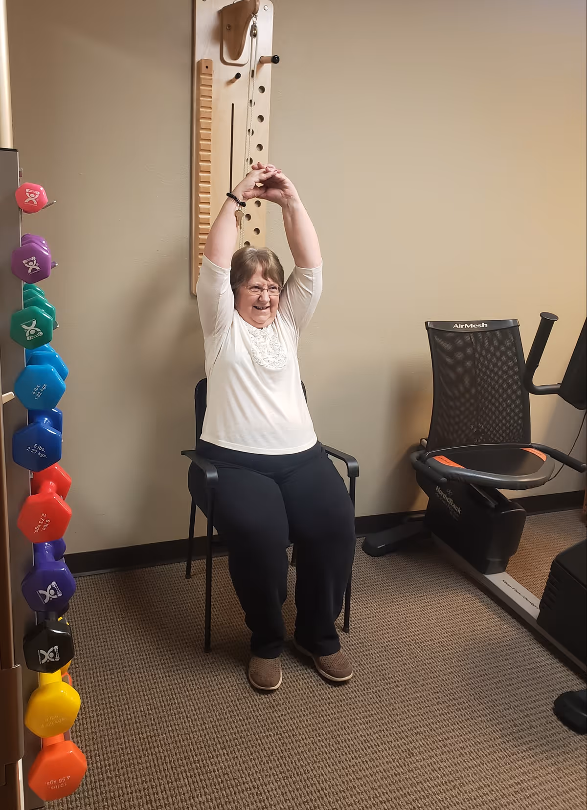 An elderly woman sitting on a chair in a fitness room, stretching her arms above her head. To her left is a rack with colorful dumbbells, and to her right is an exercise bike. The room has beige walls and carpeted flooring.