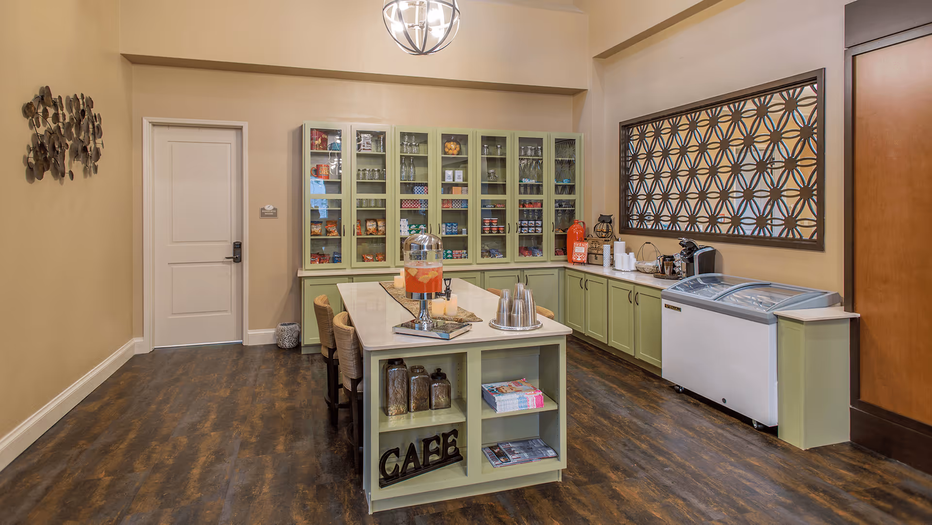 Interior view of a small cafe area in a senior living facility with green cabinets and shelves stocked with snacks and drinks. A central island has chairs, a beverage dispenser, and metal cups. There is a freezer on the right side and a decorative wall panel above the counter.