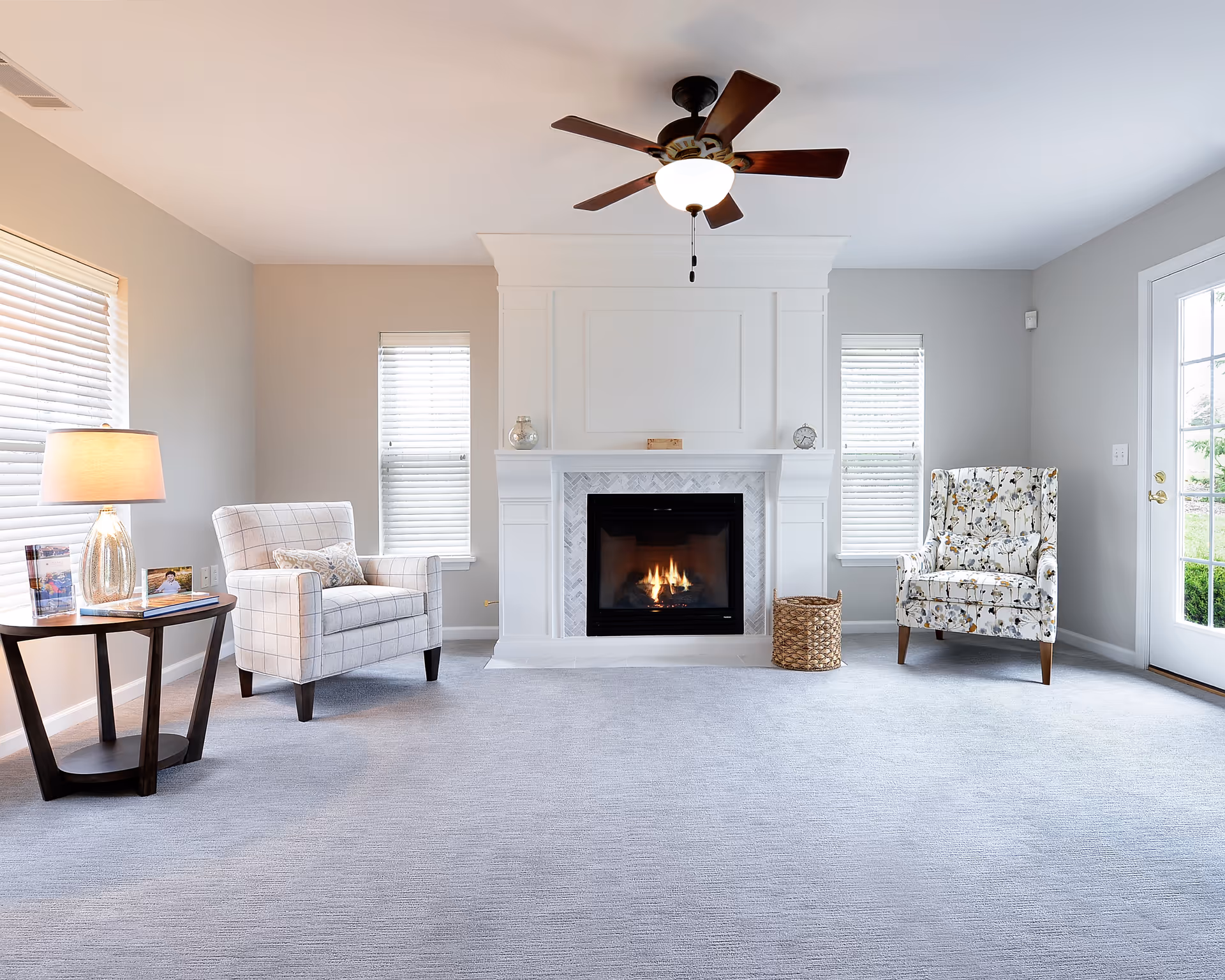 Bright, spacious living room with a central white fireplace (lit), two armchairs, a side table with lamp, ceiling fan, and a glass door to the right.