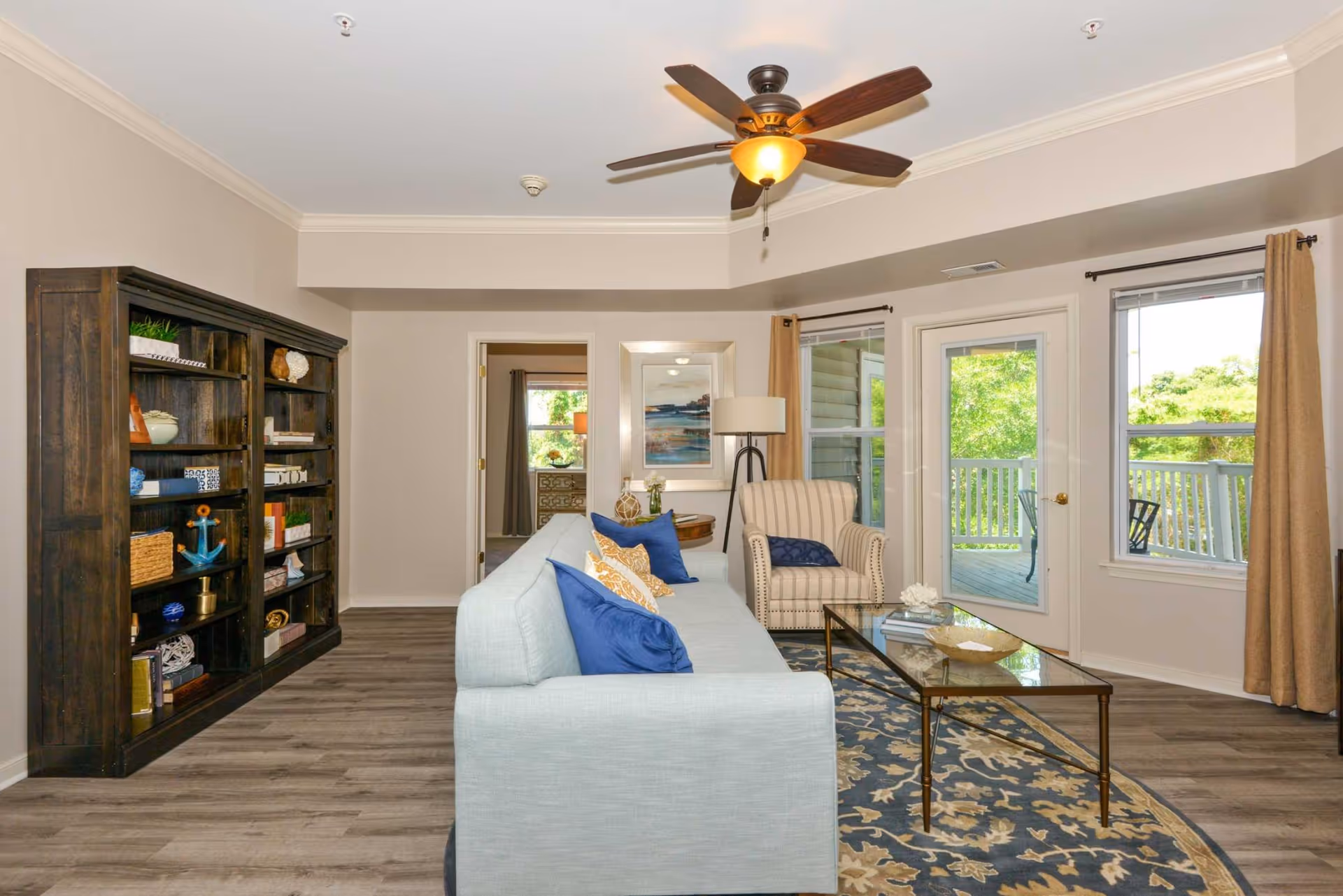 Bright living room with a light blue sofa, bookshelf, striped armchair, glass coffee table, ceiling fan, and a door leading to a balcony.