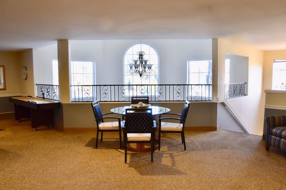 A well-lit interior common area with a round glass table surrounded by four chairs. Behind the table is a decorative wrought iron railing and three large windows, one of which is arched with a chandelier hanging in front of it. To the left, there is a shuffleboard table, and to the right, part of a striped upholstered armchair is visible. The space has beige carpet and light-colored walls.