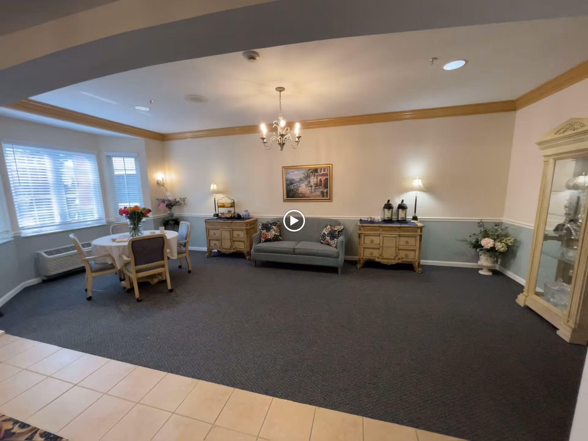 Communal sitting area with a couch and side tables centered under a chandelier, a small dining table by the windows, and a display cabinet.